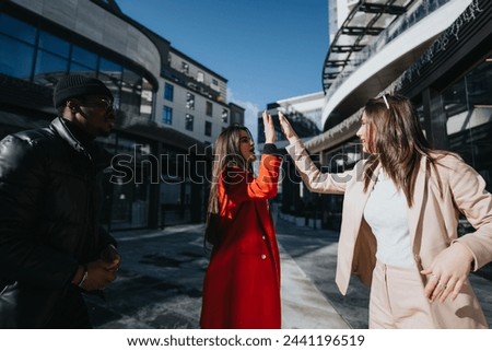 Two joyful women high-fiving with a smiling man watching, exemplifying friendship and success on a sunny city street. Royalty-Free Stock Photo #2441196519