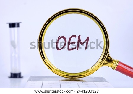 OEM original equipment manufacturer concept. Text through a magnifying glass on a gray background next to an hourglass