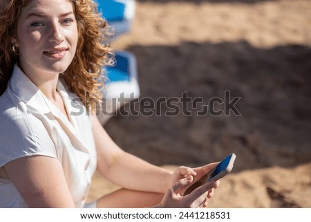 A woman is sitting on the beach and looking at her phone. She is smiling and she is enjoying herself