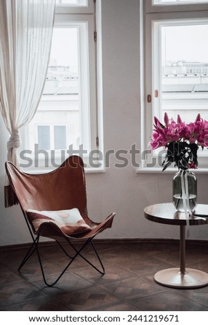Cozy corner with a leather chair and round table, highlighted by a vase of pink lilies, in a light-filled room with large windows.