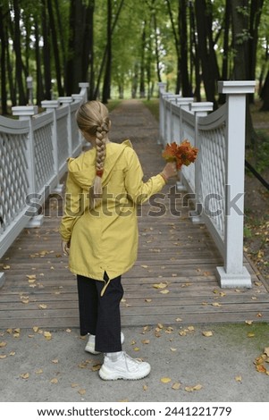 A girl with a long braid in white sneakers walks along a beautiful bridge with openwork railings in an autumn park, holding a bouquet of yellow leaves.