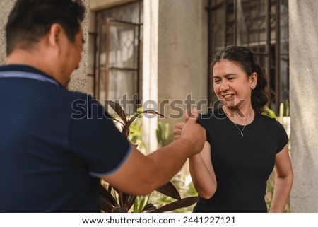 A middle-aged Southeast Asian couple shares a moment of joy while engaging in a playful game of thumb wrestling outside their house.