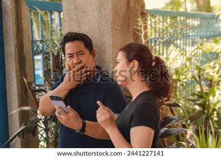 Uninterested husband yawns as he looks at his phone, not listening to wife talking animatedly outside their home.