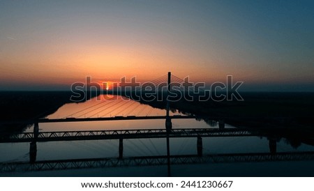 This captivating image features a serene sunset scene, showcasing the silhouette of a majestic cable-stayed bridge extending over a tranquil river. The setting sun dips below the horizon. Royalty-Free Stock Photo #2441230667