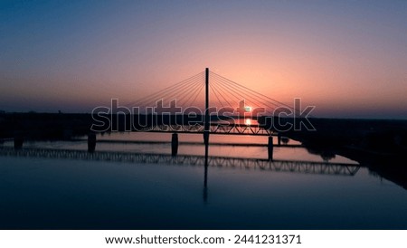 This captivating image features a serene sunset scene, showcasing the silhouette of a majestic cable-stayed bridge extending over a tranquil river. The setting sun dips below the horizon. Royalty-Free Stock Photo #2441231371
