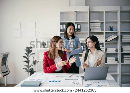 Three women are smiling and talking in a business setting. They are dressed in business attire and are standing in front of a white desk with a laptop. The room is filled with bookshelves Royalty-Free Stock Photo #2441252303