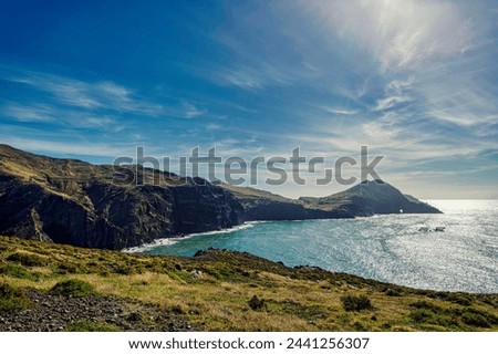 The photo depicts the picturesque landscape of Madeira PR8 Vereda da Ponta de São Lourenço: light blue sea, rocky coastline, and green hills under a blue sky.