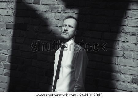 Film noir concept. Fashionable charismatic mature man stands near brick wall and looking at camera. Graphic deep shadows. Dutch angle. Text space. Monochrome outdoor shot Royalty-Free Stock Photo #2441302845
