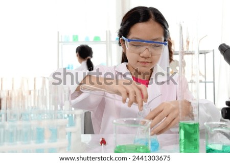 Cute young scientist schoolgirl wearing lab coat and safety glasses, doing science experiments. Student girl child using lab equipment to study chemistry in laboratory. Kid learning science education.