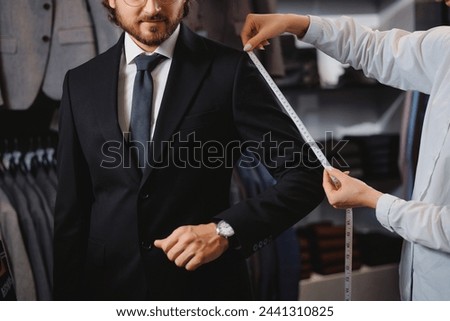 Professional dressmaker taking measurements of businessman in atelier classic menswear. Tailor fitting bespoke suit to handsome man.