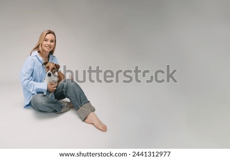 Smiling blonde woman in a blue shirt sits on the floor, the dog sitting on her laps. Funny cute friends studio portrait. Grey background. Large horizontal size banner copy empty space