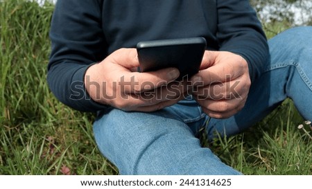 closeup of a man dressed in jeans and and blue sweatshirt using his smartphone sitting on the grass in spring