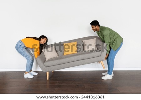 Happy indian spouses moving grey sofa in room over white wall background at their new home, moving to new apartment, full length, free space Royalty-Free Stock Photo #2441316361