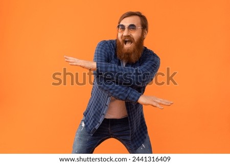 Positive redhaired hipster man with beard dancing making funny moves, exuding cool vibes and joy, partying over orange studio backdrop, wearing casual checkered shirt and jeans