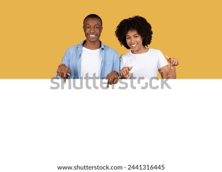 A happy millennial African American couple is pointing towards the camera with bright smiles, wearing casual clothes on a vibrant yellow background, suggesting selection or promotion