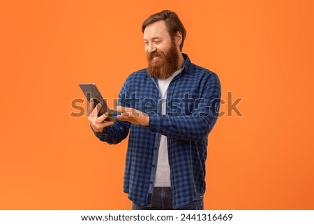 Redhaired bearded man websurfing on digital tablet device, browsing internet and communicating online through video calls, standing with gadget computer on orange studio background Royalty-Free Stock Photo #2441316469