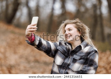 A cheerful young woman with dental braces is take a picture with her smartphone during the fall season in a park.
