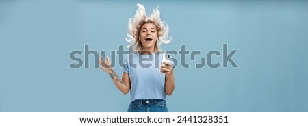 Expressing happiness with help of great tunes. Joyful amused and happy good-looking young female student jumping having fun listenign music in wireless earbuds, holding smartphone over blue wall.