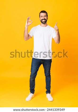 Full-length shot of man with beard over isolated yellow background showing ok sign and thumb up gesture