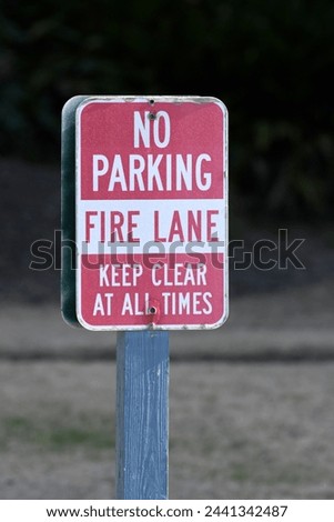 A red and white "No Parking" sign that states, "Fire Lane", "Keep Clear At All Times. The sign and sign post have a dark background in the picture.