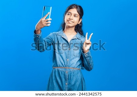 indian teenage girl taking self portrait on her mobile phone posing v- sign hand gesture with a toothy smile against a single color background