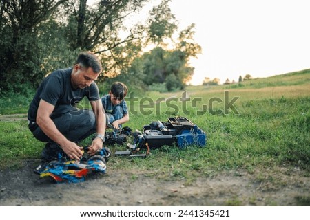 Picture of a father and his son fixing their toy race cars in nature. They have an open toolbox on the ground and they took apart their cars, to replace fresh batteries. Quality family playtime.