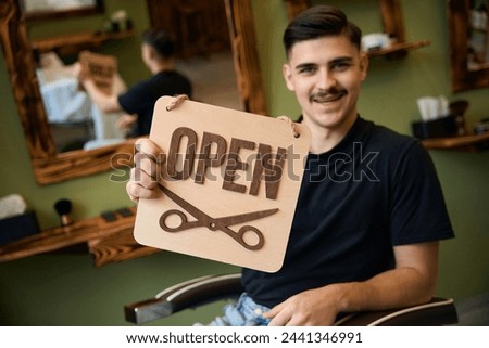 Smiling man coiffeur holding sign with open inscription in barbershop