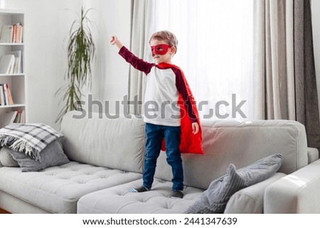 Child in superhero costume with red cape and mask standing on a sofa Royalty-Free Stock Photo #2441347639