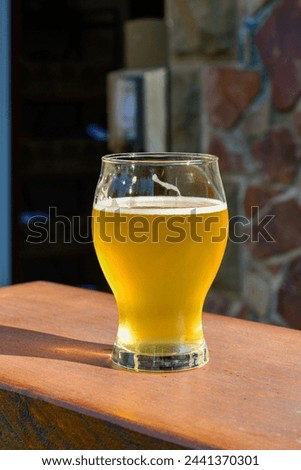 A side view of a small sample glass of pale ale craft beer. The liquid has a yellow tint. A clear beer glass sits on the edge of a wooden table at a microbrewery. The background has a rock wall.