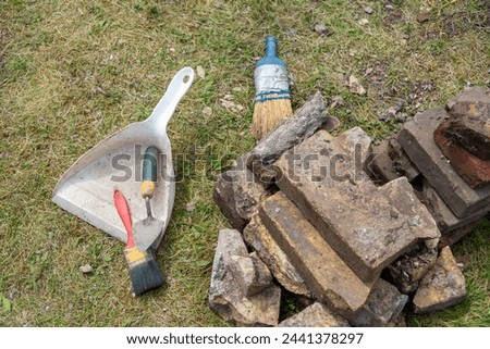 Archaeology dig tools for removing dirt and debris at a burial site. The soft bristled brushes, dust pan, and pointy trowel lay on the ground next to vintage brown bricks. There's debris on the grass. Royalty-Free Stock Photo #2441378297
