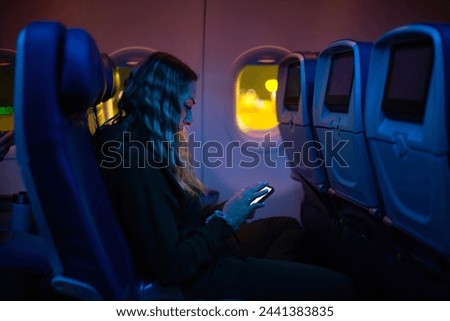 Woman passenger sitting in her seat on an airplane and browsing the internet on her mobile phone while traveling on a red-eye overnight flight Royalty-Free Stock Photo #2441383835