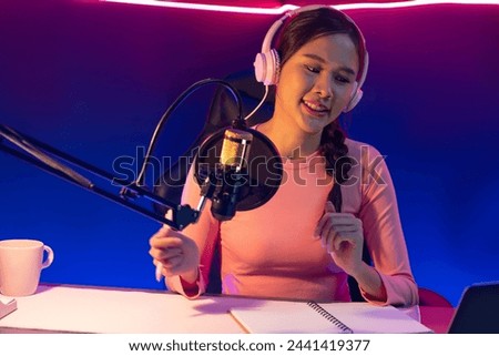 Smiling creative young beautiful Asian influencer talking in channel with positive podcast speech at night time, using laptop for presentation online at pink neon light modern studio. Stratagem.