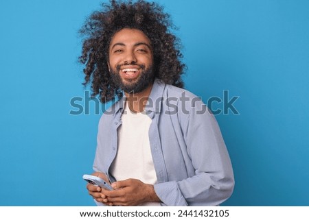 Young carefree Arabian man student laughing holding mobile phone and using instant messengers to chat with girlfriend or exchange funny pictures with college buddies stands on blue background. Royalty-Free Stock Photo #2441432105