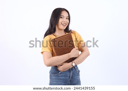 happy young Asian woman with long hair hugging book with velvet cover wearing yellow t-shirt isolated white background Royalty-Free Stock Photo #2441439155