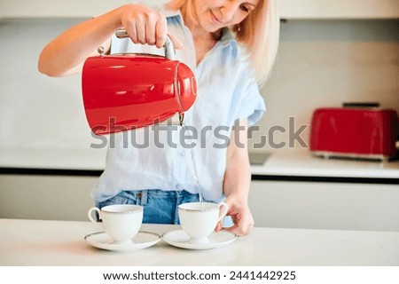 Cropped picture of woman pouring water into cup. Young female making tea for two. Woman holding red kettle over two cups.