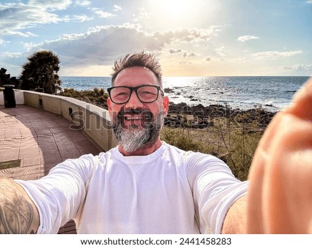 One youthful and cheerful mature man taking selfie picture with app smartphone camera smiling and enjoying outdoor leisure activity on vacation summer holiday with ocean beach and blue sky background