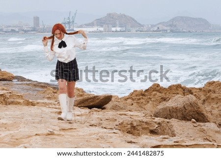 Senior woman playing pretend in a school uniform while walking on the beach and pulling her red hair in pigtails. Royalty-Free Stock Photo #2441482875