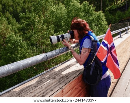 Female tourist with norwegian flag taking travel picture from Stegastein viewing platform. National tourist route Aurlandsfjellet.