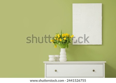 Beautiful vase of yellow tulips on chest of drawers and picture on wall in living room