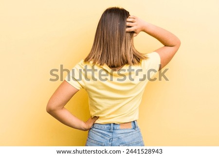 pretty hispanic woman feeling clueless and confused, thinking a solution, with hand on hip and other on head, rear view