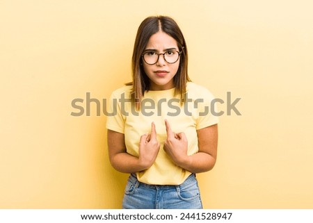 pretty hispanic woman pointing to self with a confused and quizzical look, shocked and surprised to be chosen