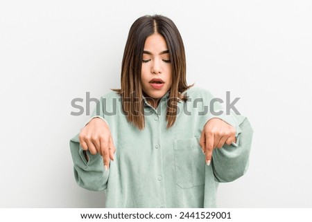 pretty hispanic woman with open mouth pointing downwards with both hands, looking shocked, amazed and surprised
