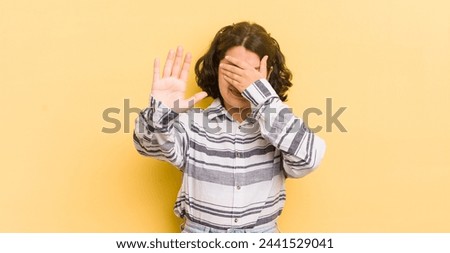 pretty hispanic woman covering face with hand and putting other hand up front to stop camera, refusing photos or pictures