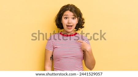 pretty hispanic woman looking shocked and surprised with mouth wide open, pointing to self