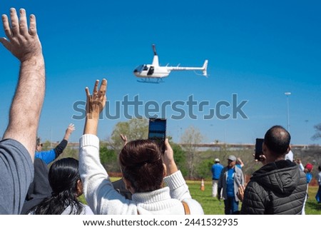 Diverse people hands waving, taking photo hovering helicopter drops colorful eggs, goodies at Easter event, Richardson, Texas, rear view crowd group with mobile phone, holiday Chistian tradition. USA