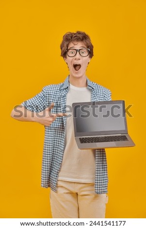 Modern education. A smart student boy wearing glasses holds a laptop in front of him and makes his eyes wide and opens his mouth in surprise. Yellow studio background. Copy space.