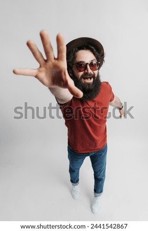 Cheerful bearded man with sunglasses and a hat reaches out towards the camera, creating a sense of interaction. His joyful expression and casual outfit convey a relaxed and playful atmosphere. Royalty-Free Stock Photo #2441542867