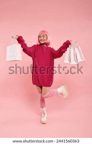 cute asian woman in her 20s posing with leg raised and holding shopping bags on pink background