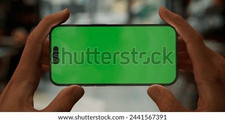 Person holding a smartphone with a green screen in a blurred cafe setting, perfect for showcasing apps and mobile technology. Horizontal phone orientation