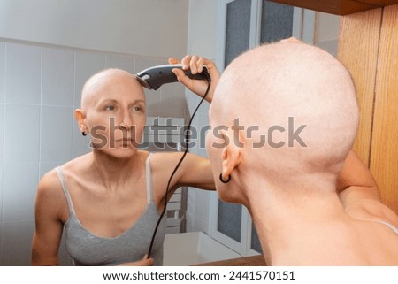 A bald woman is trimming her head with electric clippers while looking into the mirror Royalty-Free Stock Photo #2441570151